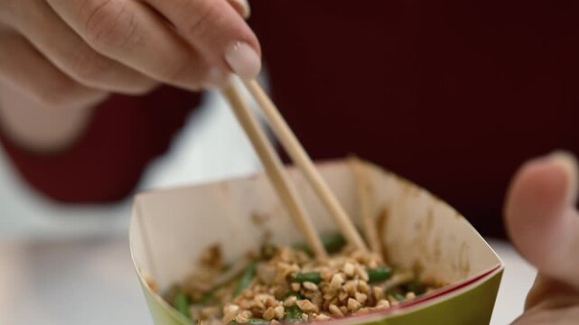 Person holding chopsticks with noodles in a takeout container during a lunch break at restaurant