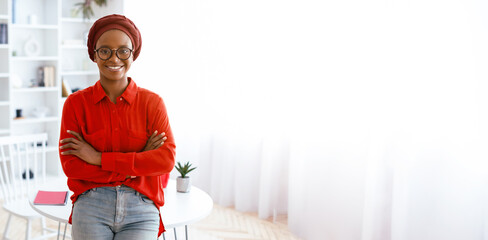 Woman with glasses stands with arms crossed in a bright room. She wears a red top and a headwrap. The setting includes plants and light furniture. Natural light fills the space © Prostock-studio