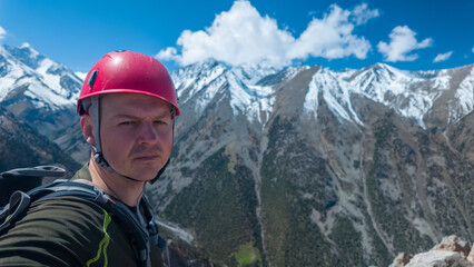 A man stands on a mountain peak taking a selfie. The sky is clear with some clouds. Snow-covered mountains surround him. He wears climbing gear and a red helmet. © Evgeniy