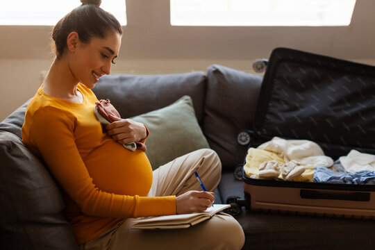 Expectant mother sitting with baby clothes near open suitcase, writing preparation list