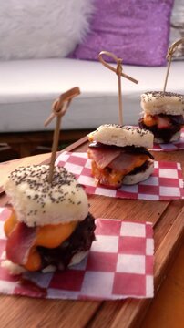 Close up tracking shot of gourmet beef sliders with cheddar and bacon served on wooden board at elegant outdoor park catering event