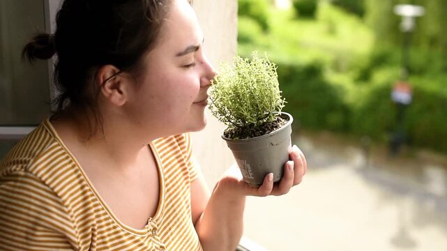 A woman smells a fresh green thyme plant in a plastic pot with French recycling text near a window. She holds the aromatic herbal container during an eco friendly activity.