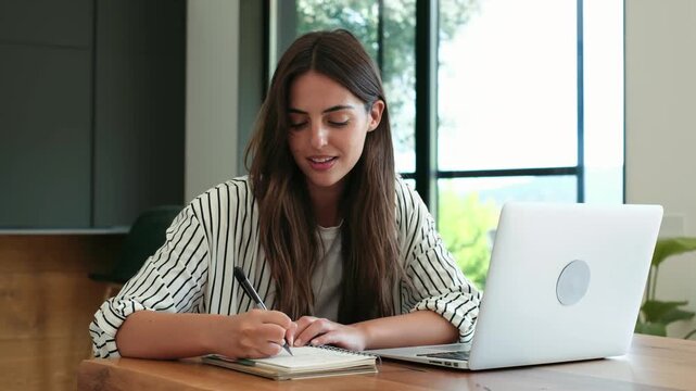 Beautiful young woman attending an online class on her laptop, smiling while taking notes in a notebook at her desk. Remote education and modern student lifestyle concept in a bright interior