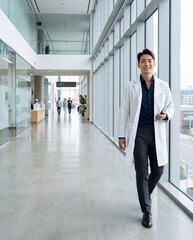 Fototapeta premium Smiling confident young Asian male doctor in white lab coat walking through modern bright hospital corridor, premium healthcare medical portrait
