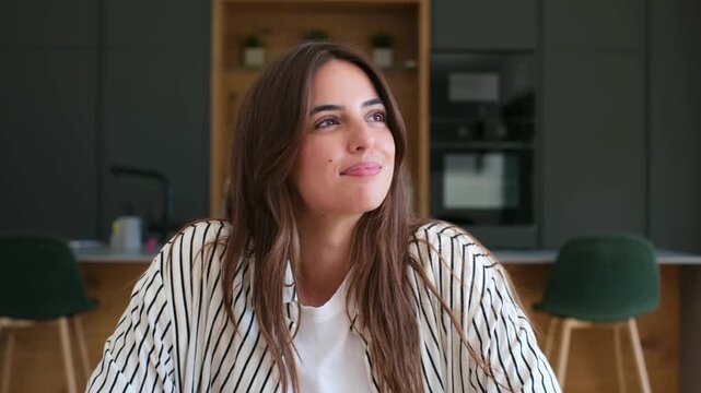 Beautiful young woman with a genuine smile looking away thoughtfully while sitting in her modern home kitchen. A charming portrait expressing positivity, happiness, and contentment in daily life