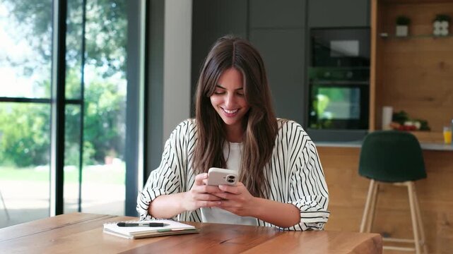 Cheerful young woman sitting at a wooden table in a modern kitchen, smiling while texting on her smartphone and enjoying a pleasant conversation on social media or a dating app