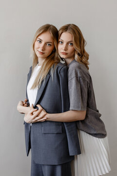 Two young women pose closely together against a plain light background, one wearing a tailored dark suit and the other in a gray top with a white pleated skirt