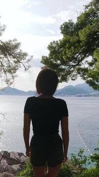Back view of female traveler walking in the park and approaching the top cliff with open arms in front of amazing seascape at sunset, outdoors. 4k