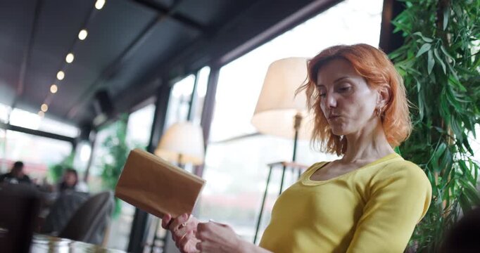 Woman opens yellow envelope at table inside cafe near window during daytime in bright yellow long sleeve shirt