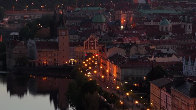 Aerial drone view of the Old Town waterfront with a clock tower, baroque domes and red rooftops reflected in the Vltava river at dusk.