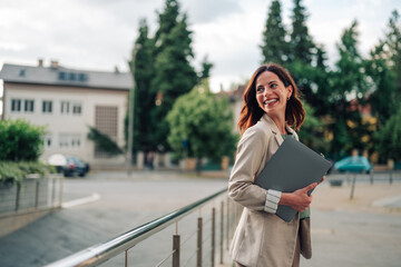 Woman with laptop smiling, looking away in urban environment, embodying business success and...