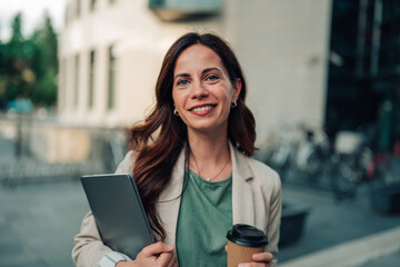 Fototapeta premium Confident businesswoman walking in urban setting holding digital tablet and takeaway coffee cup, smiling at camera