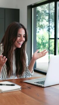 Happy businesswoman making a video conference call on her laptop computer, looking surprised and celebrating good news while sitting at a desk in her modern home office near a window