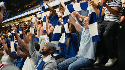 Excited blue team football supporters raising arms and cheering in packed stadium stands during thrilling live soccer match atmosphere, Soccer fans shouting and singing chants, global event. © DC Studio
