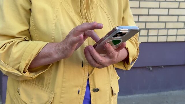 Close-up view of an older persons midsection as they hold a small silver point-and-shoot camera. The subject wears a yellow lightweight jacket with buttons