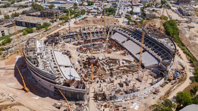 Large construction site actively building a new stadium, featuring multiple cranes lifting materials and ongoing development of concrete structures for the future sports and entertainment arena