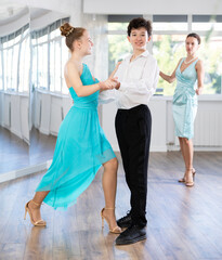 Naklejka premium Positive teenage boy and girl in performance outfit confidently performing tango steps at dance rehearsal in choreography studio while female teacher observing from background..
