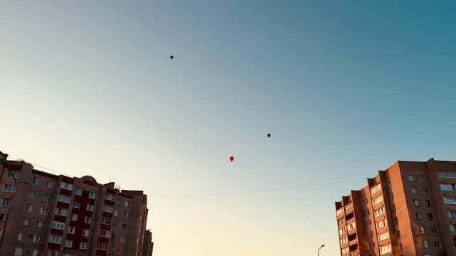 Low-angle dusk view between two apartment blocks showing a small cluster of colorful balloons floating above rooftop level against a blue-to-amber gradient