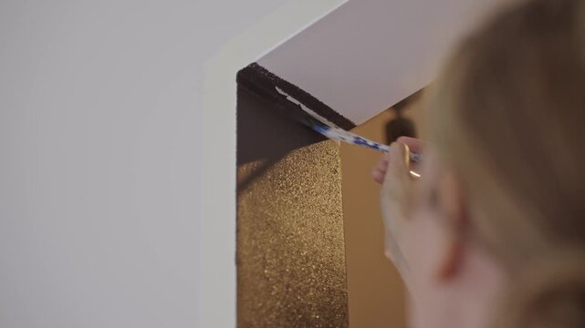 Woman applies dark paint along doorway edge with brush near white wall