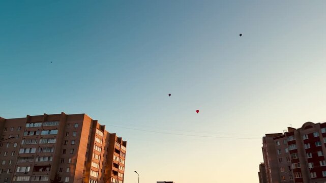 Low-angle dusk view between two apartment blocks showing a small cluster of colorful balloons floating above rooftop level against a blue-to-amber gradient