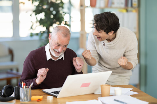 Happy young and elderly men colleagues celebrating victory or positive result while looking at laptop screen in office