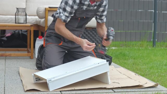 Man inserts screw and fastens white drawer unit with cordless drill on cardboard outdoors