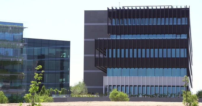 Contemporary urban architecture featuring two modern buildings with glass facades, steel structures and landscaped greenery in the foreground. Static exterior view under clear daylight with clean geom