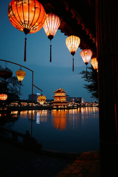 Lanterns glow by a lake with buildings in the evening light