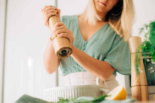 Woman seasoning raw chicken with wooden pepper mill in bright ki