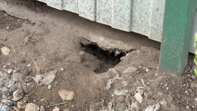 Close-up view of a corrugated metal exterior wall with green painted trim beside an uneven gravel and soil foundation. Small rocks and debris scatter
