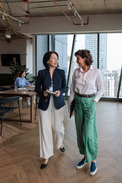 Two businesswomen converse while walking in a modern office space.