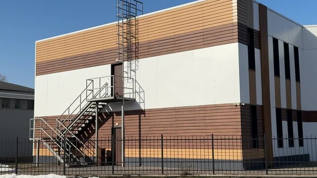 Stylish modern house with siding under blue sky, geometric facade of glass and wood panels, parking lot and metal fence