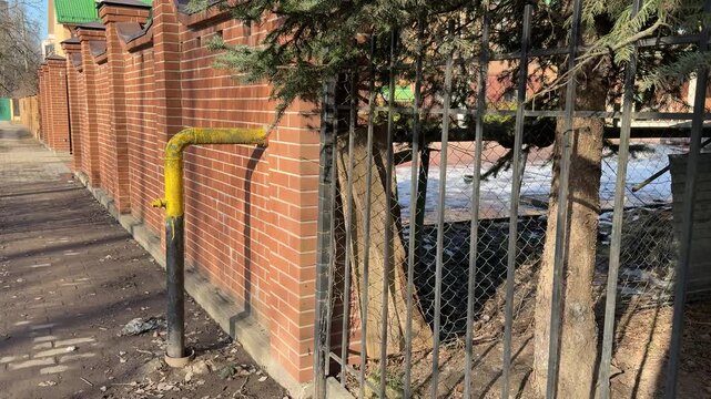 Yellow gas pipe through brick wall showing residential supply to cottage village, metal support post and iron fence, evergreen