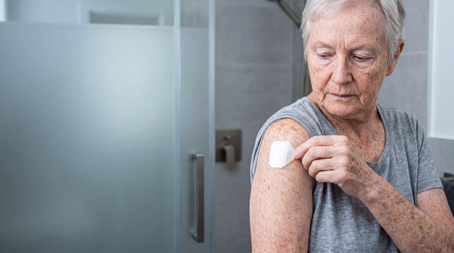 Elderly woman applying adhesive medicated patch on arm for cardiovascular disease treatment. Senior patient managing heart health condition with topical medicine. Geriatric healthcare concept.