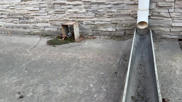 White downpipe and towed water crane sit against stone facade on paved yard, small planter with weeds and metal bracket, scattered