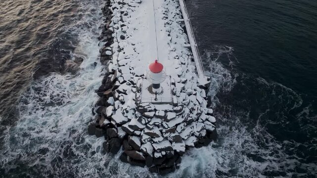 Aerial view of small lighthouse on snowy rocky breakwater in winter sea