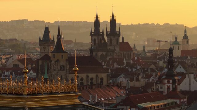 Aerial drone view of the National Theatre gilded rooftop balustrade with Old Town Square towers and Tyn Church silhouetted in warm golden sunrise haze, Prague, Czech Republic.