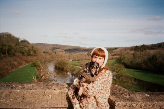 Woman Holds Dog on a Bridge by the River in a Green Valley