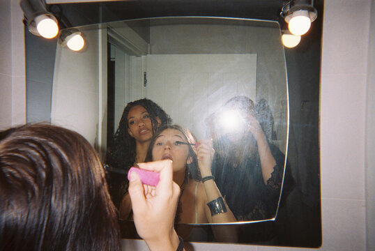 Friends applying makeup together in bathroom mirror