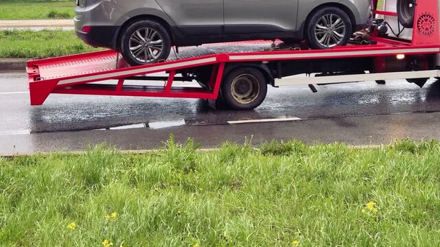 Close-up passenger car on red flatbed tow truck, spring roadside scene with wet pavement and puddles, green grassy verge dotted