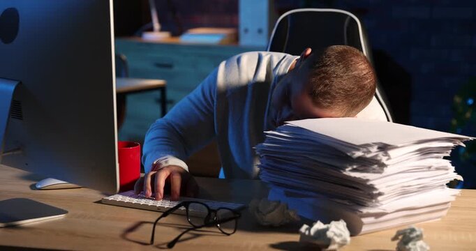 Workaholic. Tired man working with computer and leaning on stack of documents at wooden desk indoors at night. Camera moving forward