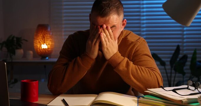 Workaholic. Overwhelmed man at wooden desk with laptop and notebook indoors at night, camera moving forward