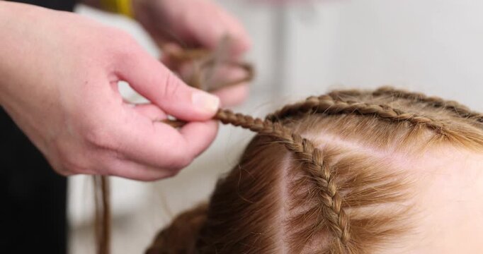 Hairstylist braiding teenage girl's hair in salon, closeup. Camera moving forward and backwards