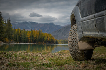 Close up of offroad SUV car tire and wheel on grass by mountain lake. 4x4 vehicle traveling through remote scenic autumn forest wilderness under dramatic cloudy sky. © ANDREY