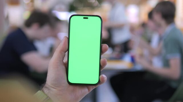 Hand holds phone with green screen. Background shows blurred people in cafe. Focus remains on phone throughout. Natural lighting illuminates scene. No animals or plants visible
