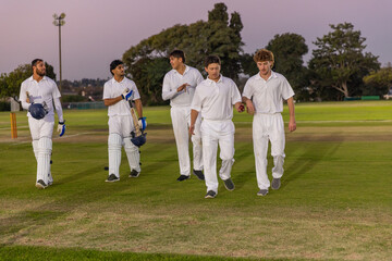 Male cricket teammates in whites walking across dusk pitch on field, carrying bats helmets and ball