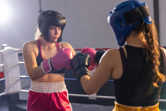 Two female boxers sparring in gym ring, wearing headgear, sports tops, pink and black gloves