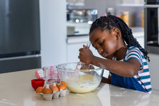 African child wearing blue apron stirring batter in clear mixing bowl at kitchen island with whisk