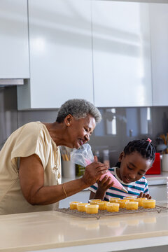 Senior grandma guiding youth granddaughter piping pink icing from clear bag on cupcakes in kitchen