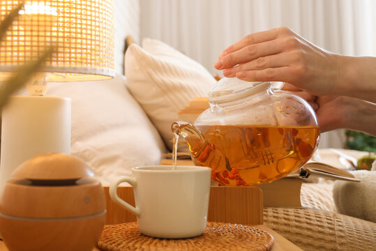 Female hands pouring green tea from teapot into cup near diffuser and lamp on bedside table in bedroom. Closeup